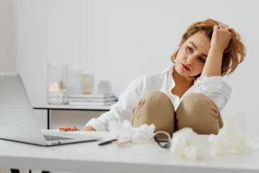 Woman appearing stressed and overwhelmed at her desk, surrounded by crumpled tissues and a laptop, highlighting the impact of stress and anxiety in daily life.