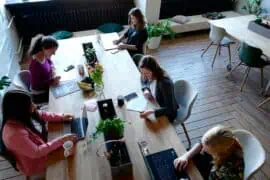 Group of women working on laptops and tablets in a bright, modern office space with wooden floors and large windows.