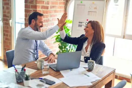 Two professionals, a man and a woman, high-fiving in a modern office setting with a laptop and documents on the table, celebrating a business achievement.