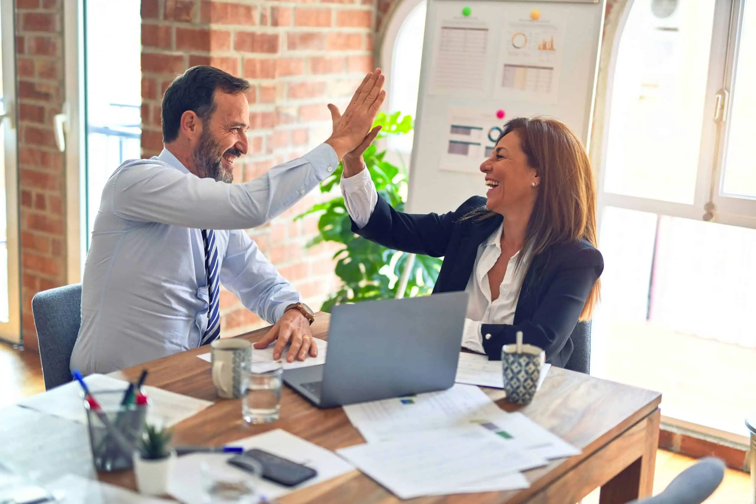 Two professionals, a man and a woman, high-fiving in a modern office setting with a laptop and documents on the table, celebrating a business achievement.