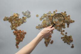Hand holding magnifying glass over a world map made of coins, symbolising global business and economic analysis.