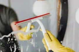 Person wearing yellow gloves cleaning a window with a squeegee, removing soap suds, in a bright indoor setting.