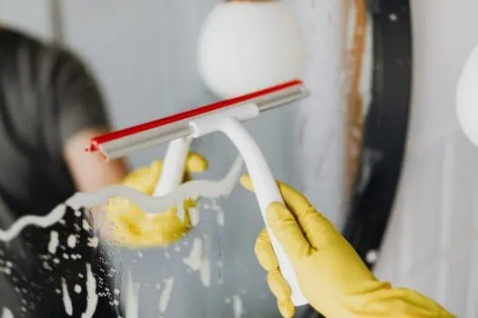 Person wearing yellow gloves cleaning a window with a squeegee, removing soap suds, in a bright indoor setting.