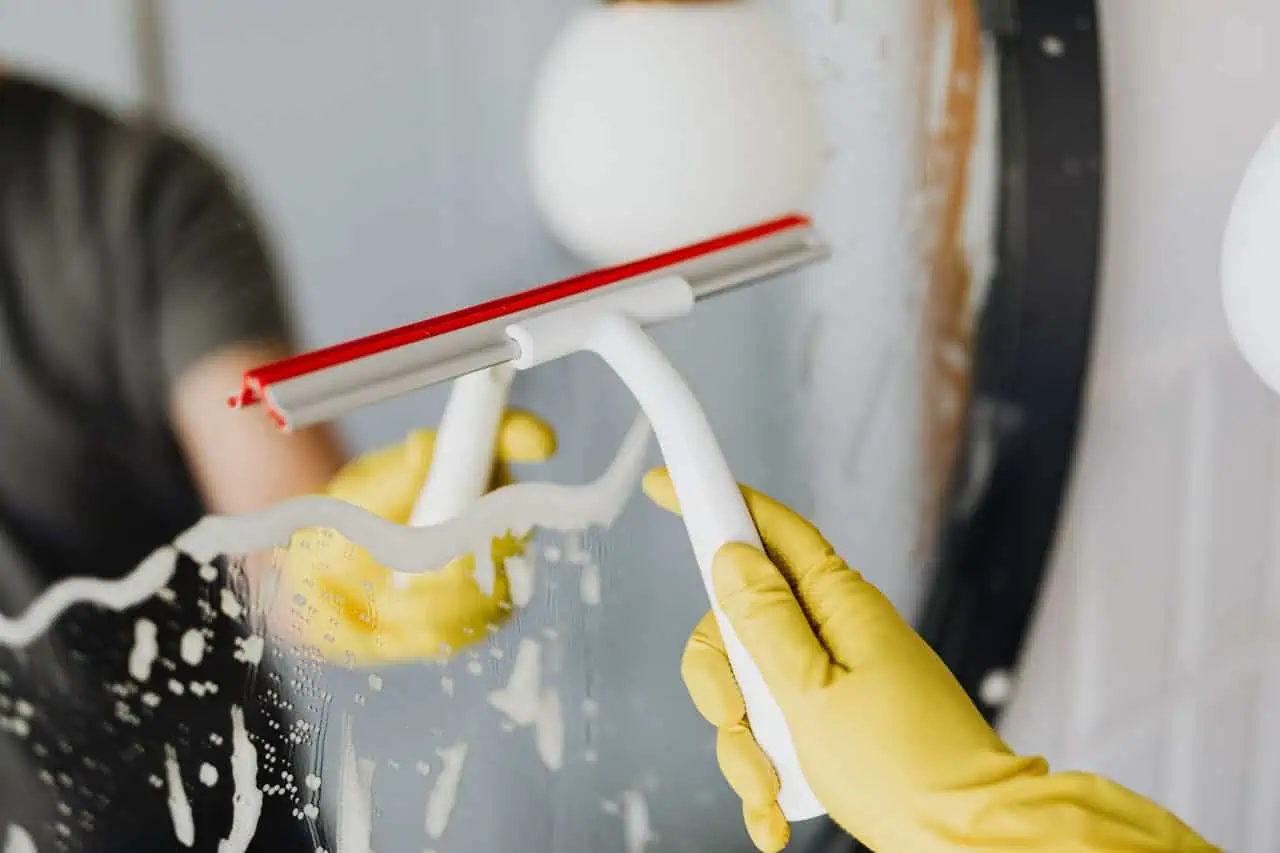 Person wearing yellow gloves cleaning a window with a squeegee, removing soap suds, in a bright indoor setting.