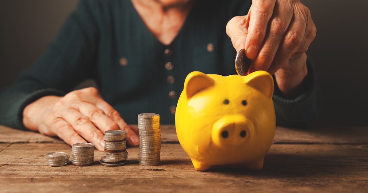 Elderly hands depositing coins into a yellow piggy bank.