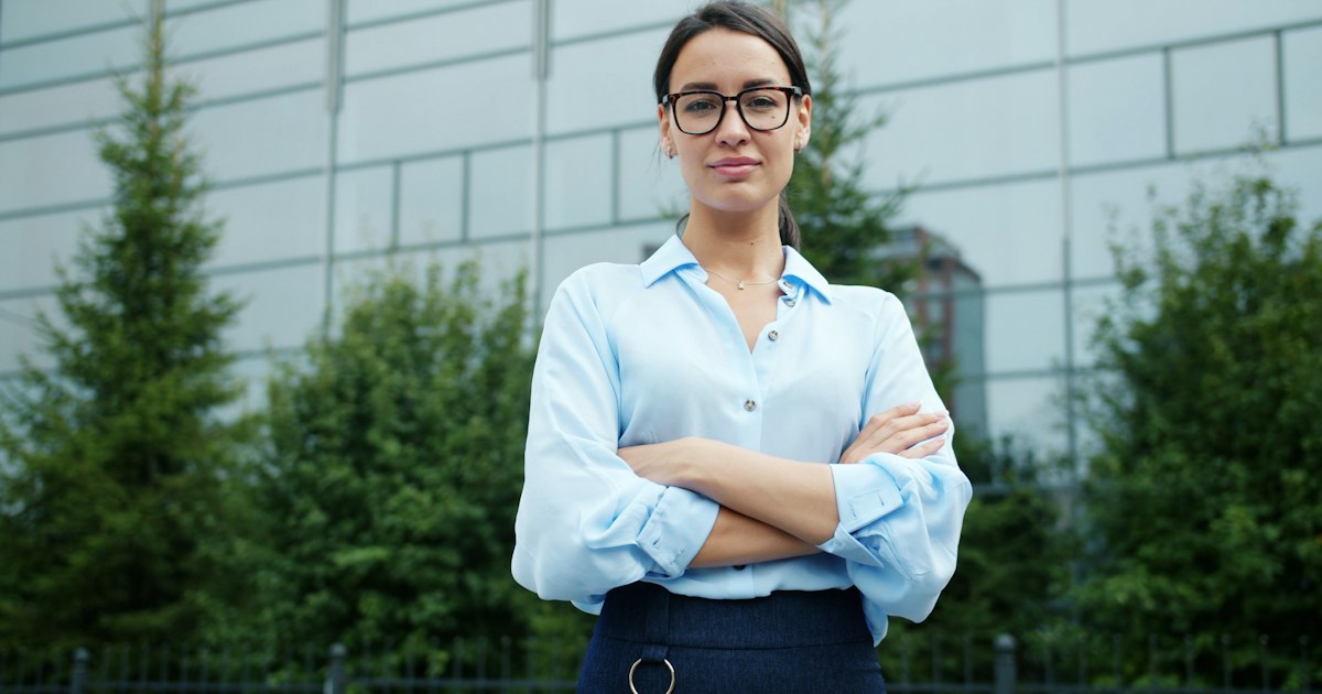 Beautiful young businesswoman in elegant formal clothing is standing outdoors with arms crossed smiling looking at camera. Female business and career concept.