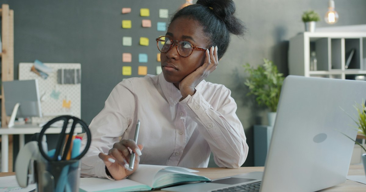Serious African American businesswoman is thinking and looking around sitting at desk in office. Businesspeople, workplace and emotions concept.