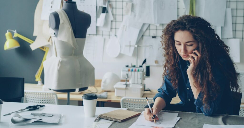 Professional female fashion designer is talking on mobile phone and drawing clothing sketch with pencil at table in tailoring shop. Everyday work and communication in workplace concept.