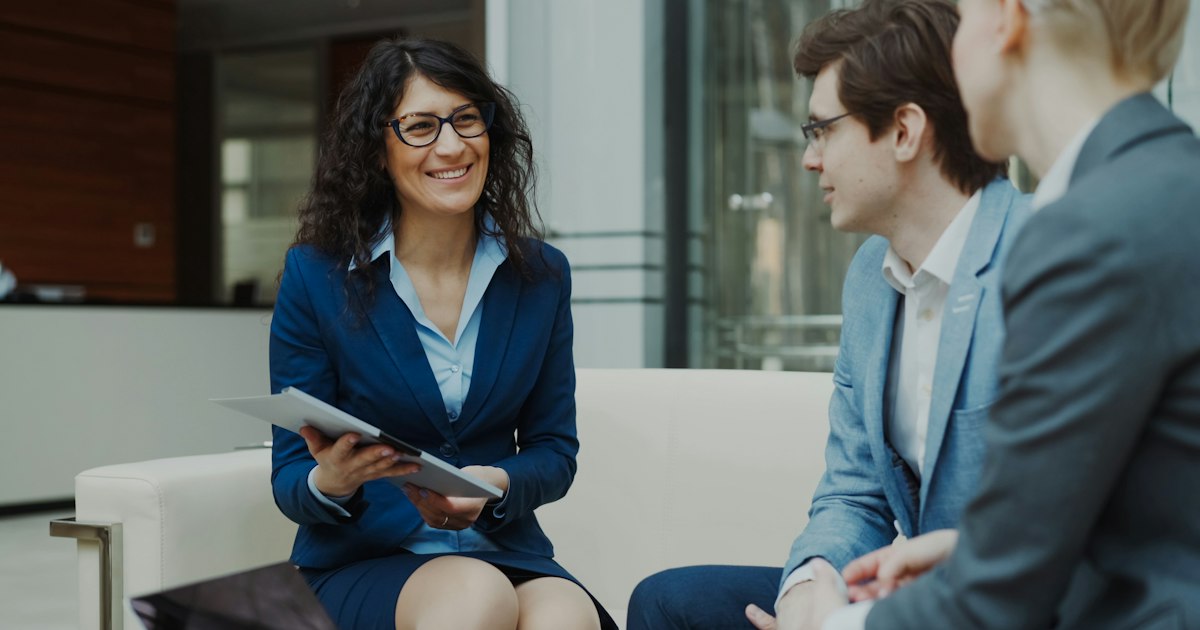 Businesswoman in glasses talking and duscussing future contract with business partners sitting on couch in modern office indoors