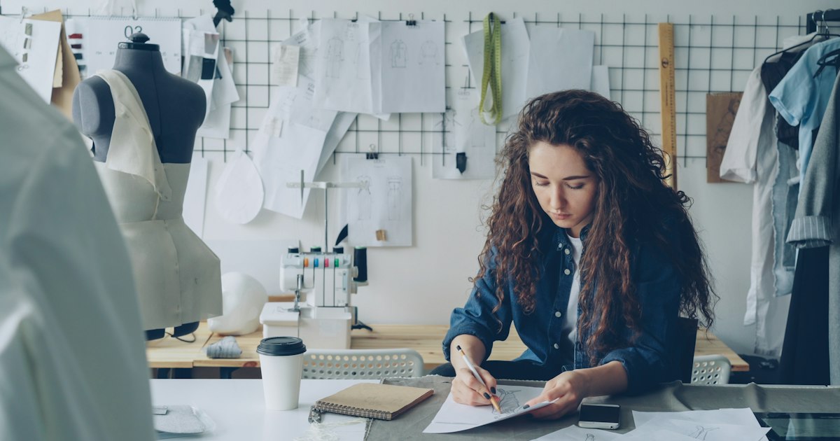 Young woman fashion designer is drawing ladies' garment sketch at desk in modern workshop. Mannequin, sewing items, to-go coffee are visible. Creativity in clothing manufacturing concept.
