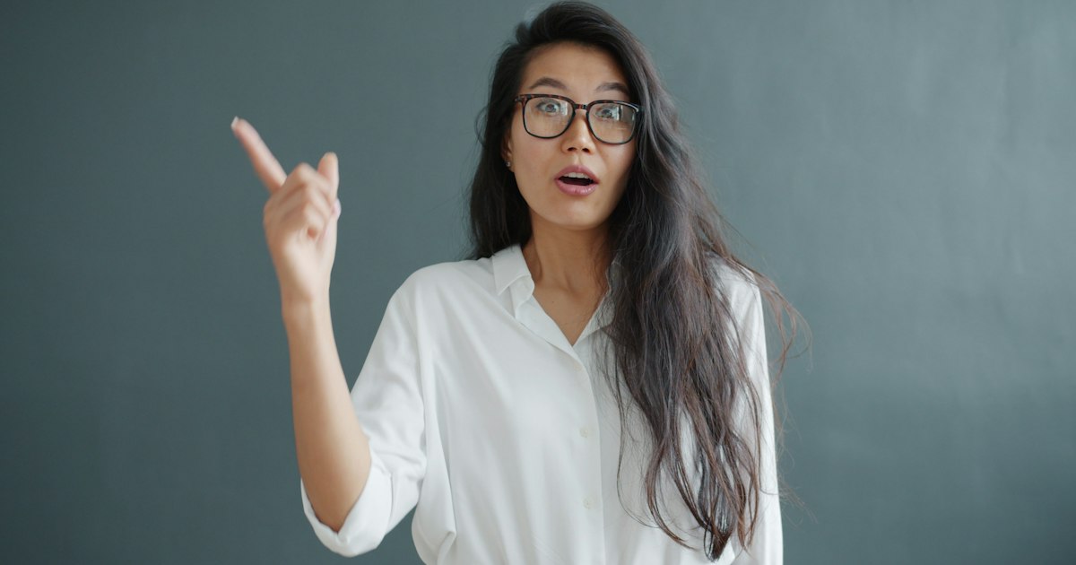smart Asian girl in eyeglasses raising finger having great idea on gray background. Successful creative people and good solutions concept.