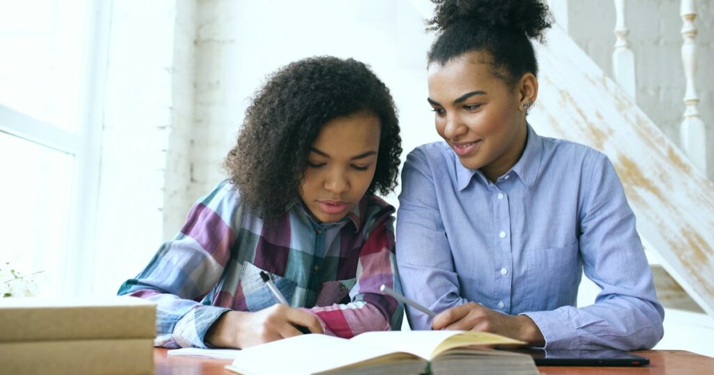 Teenage curly haired mixed race young girl sitting at the table concentrating focused learning lessons and her elder sister helps her studying at home