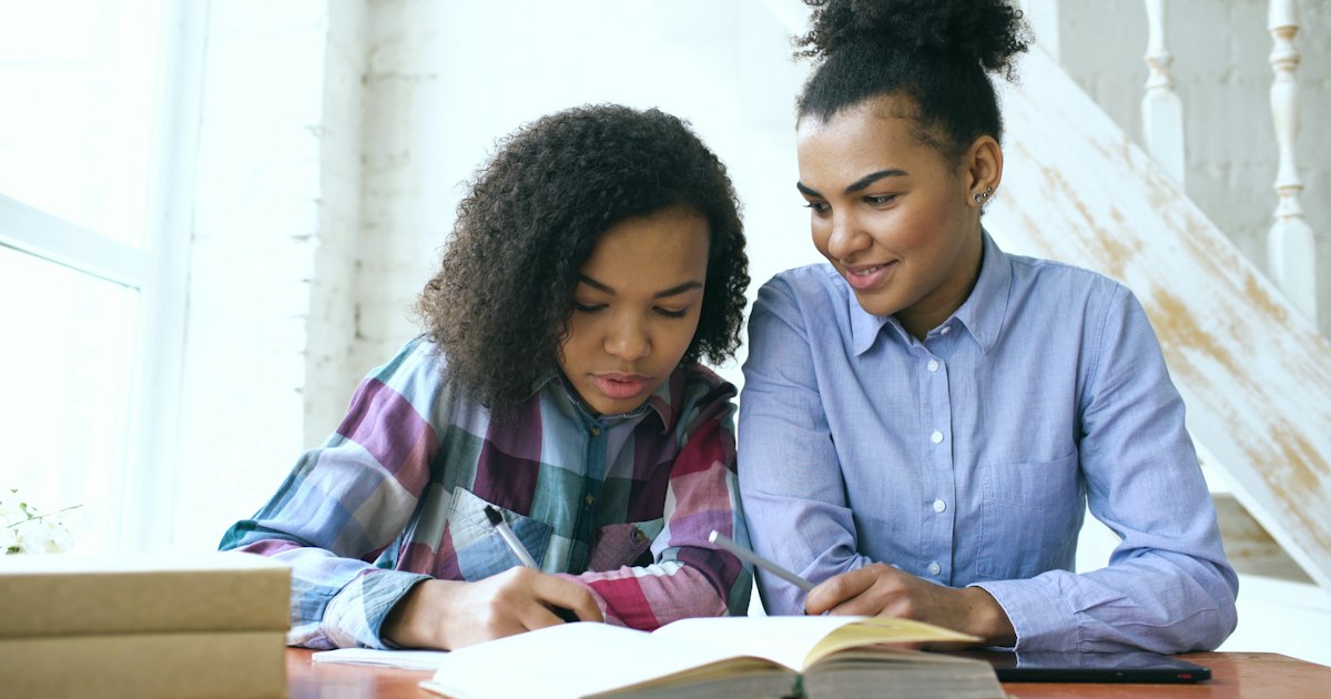Teenage curly haired mixed race young girl sitting at the table concentrating focused learning lessons and her elder sister helps her studying at home