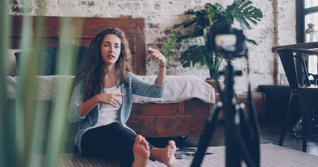 Emotional young woman vlogger is talking and gesturing recording video with professional camera for internet blog. Girl is wearing casual clothing and sitting on bedroom floor at home.