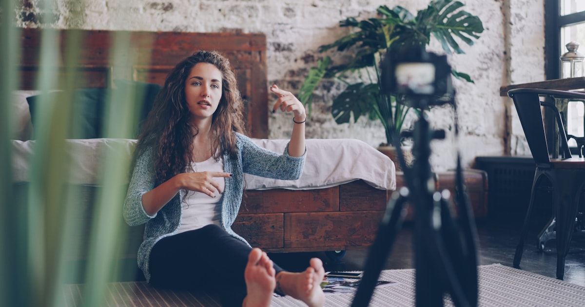 Emotional young woman vlogger is talking and gesturing recording video with professional camera for internet blog. Girl is wearing casual clothing and sitting on bedroom floor at home.