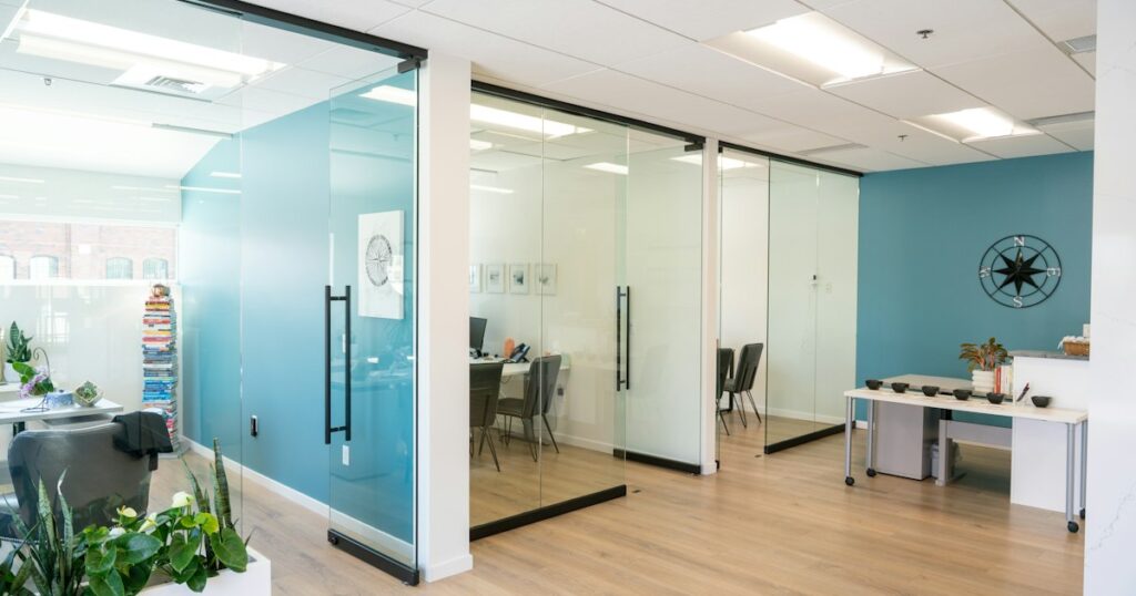 Glass-walled offices line the hallway at Deliberate Directions, reflecting a clean, open layout that supports transparency and focused work. Teal accent walls, light wood floors, and touches of greenery create a calm and modern environment. A compass wall piece and stack of leadership books reinforce the firm's coaching philosophy: clarity, direction, and continuous growth.