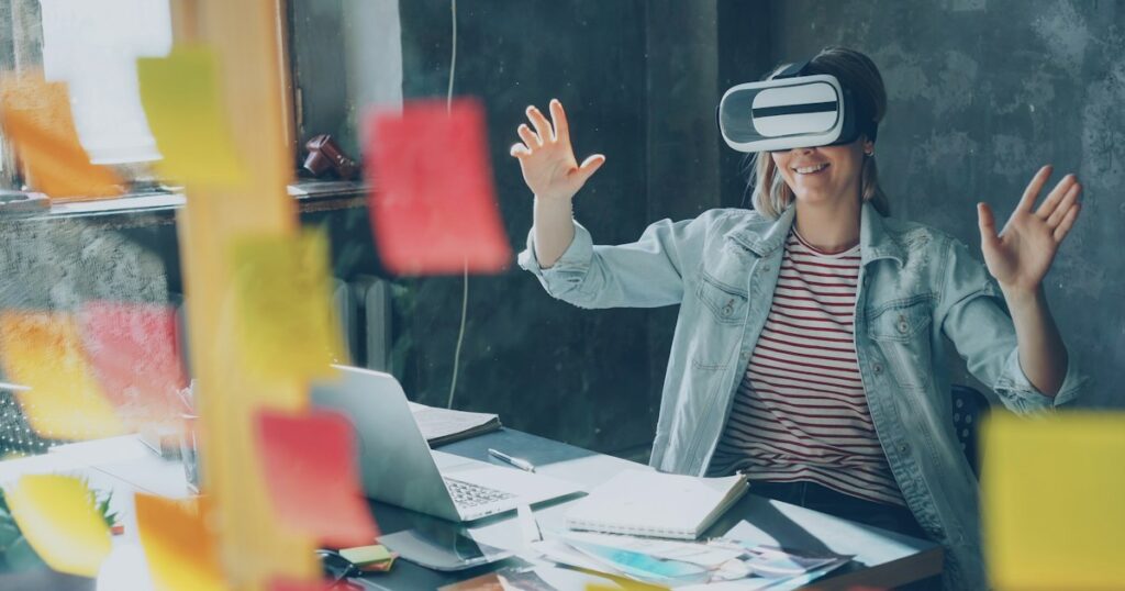 Attractive young woman is sitting at table and wearing virtual reality glasses. She is moving hands and and having VR experience in modern lof office indoors