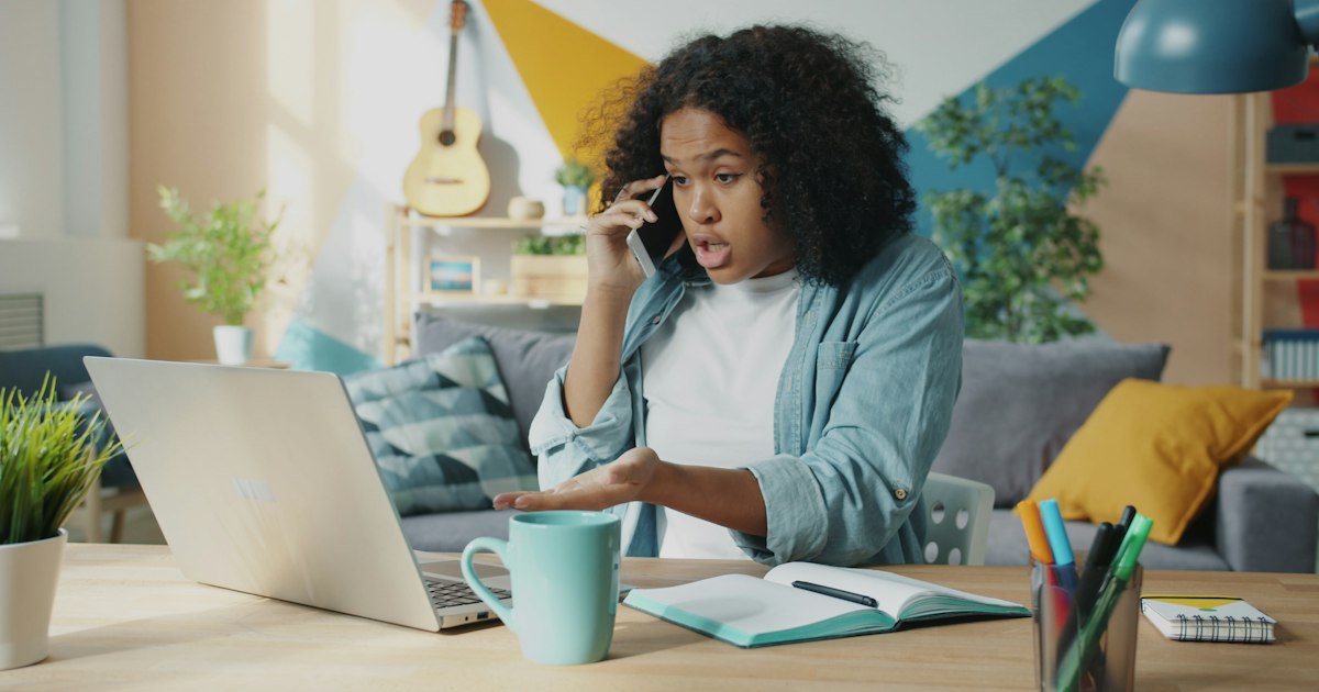 Mad Afro-American woman is yelling on mobile phone using laptop at home working online doing freelance work. Negative emotions and communication concept.
